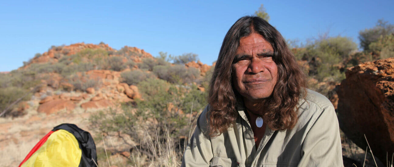 An Indigenous Elder sitting in the outback with the Aboriginal flag in the back ground