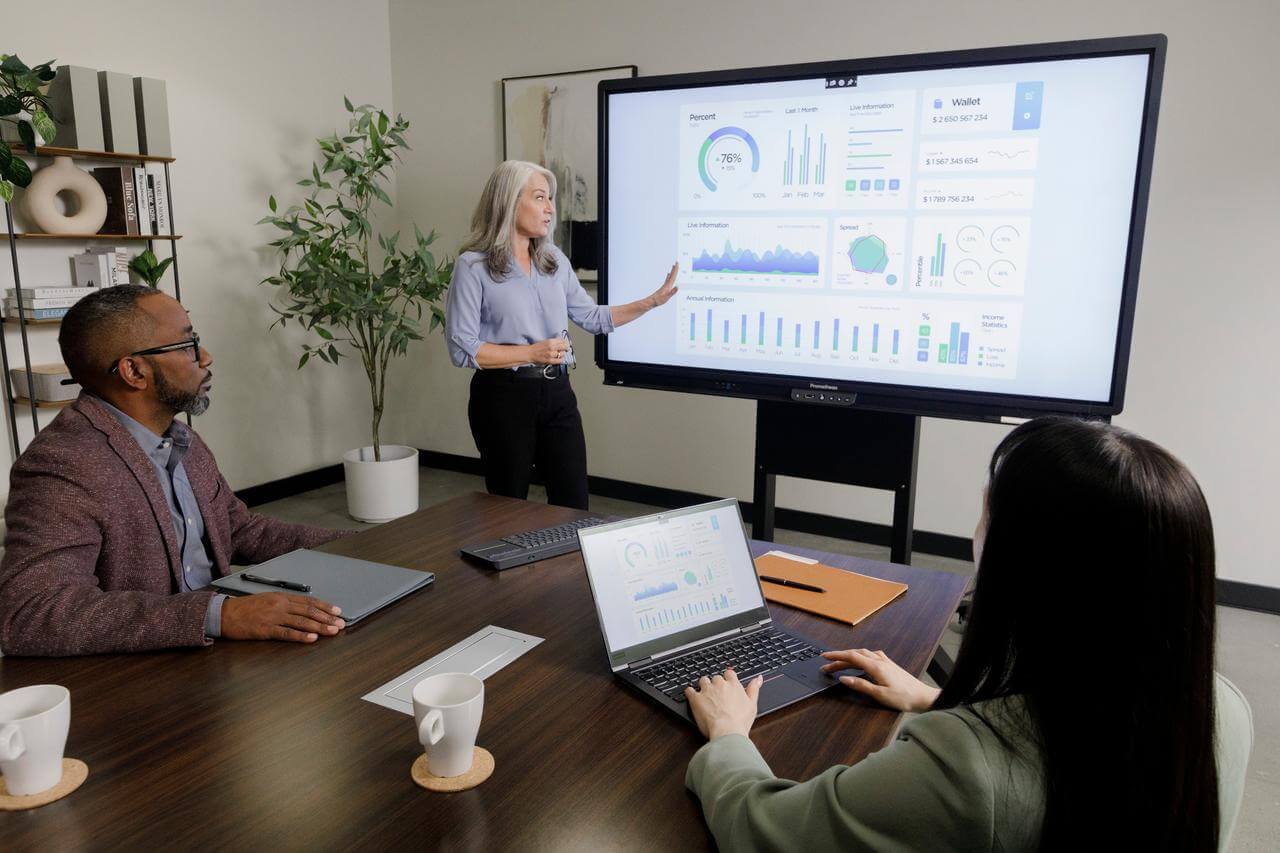 A team of professionals sat around a table in a conference room watching a woman present using an interactive display.