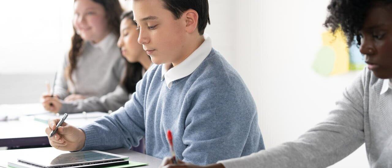 In a classroom setting, one student uses a tablet for note-taking, while others nearby write in notebooks with pens.