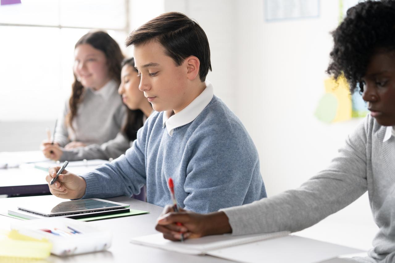 In a classroom setting, one student uses a tablet for note-taking, while others nearby write in notebooks with pens.